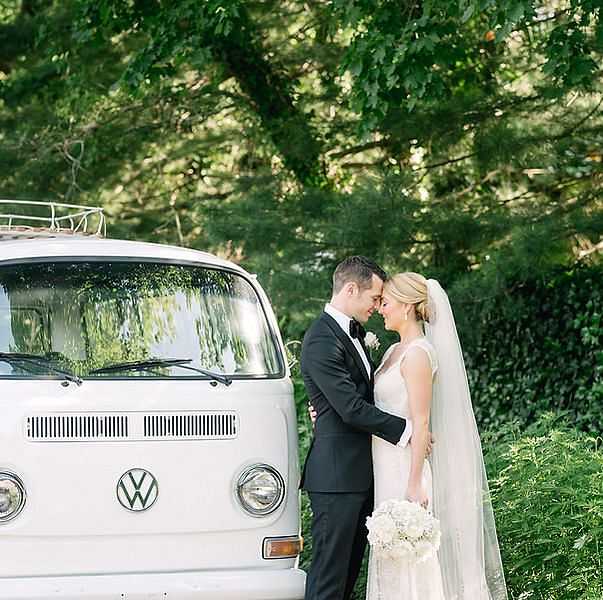 Bride and groom embrace in front of a white van in a lush green setting.