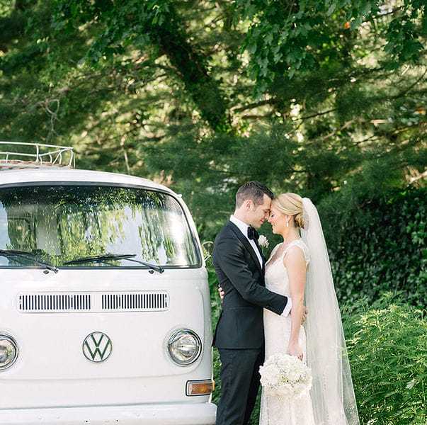 Bride and groom embrace in front of a white van in a lush green setting.